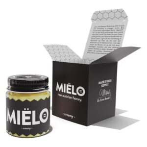 Ein Glas mit cremigem, rohem österreichischen Honig in schwarzer Verpackung mit einem offenen Deckel und beschreibendem Inhalt.